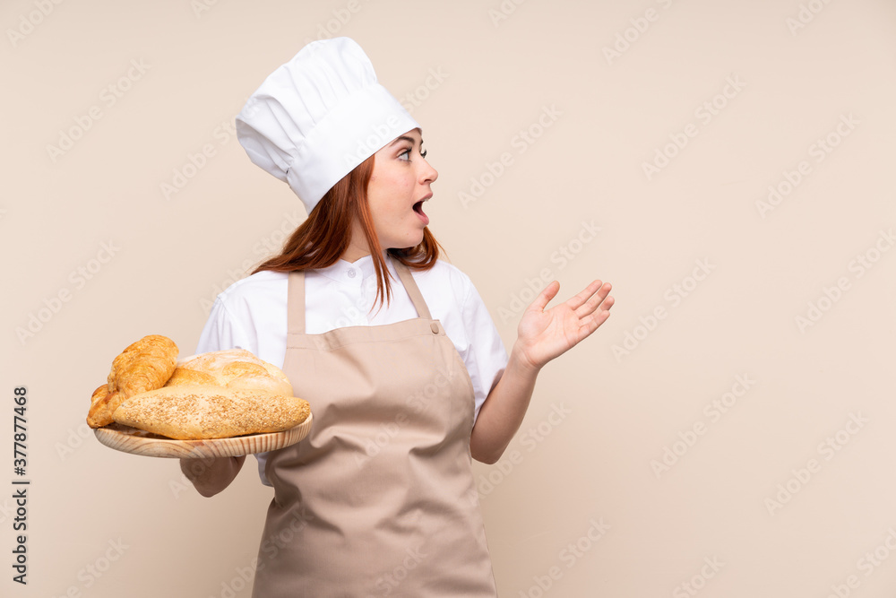 Redhead teenager girl in chef uniform. Female baker holding a table with several breads with surprise facial expression Redhead teenager girl in chef uniform. Female baker holding a table with several breads with surprise facial expression