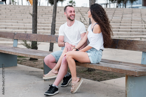 Young couple chatting and talking sitting on a bench at an outdoor park. Romantic and sweet boyfriend and girlfriend having a good time and smiling