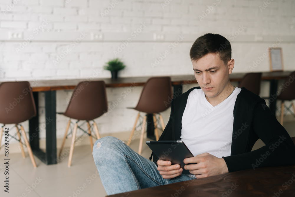 Handsome young man wearing casual fashion clothes sits at table and uses tablet. Relaxed freelancer working from home office sitting at a table with digital tablet.