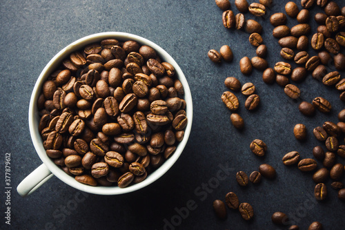 Coffee mug top view. White ceramic cup filled with coffee beans. Black table surface with coffee seeds. Morning caffeine background.