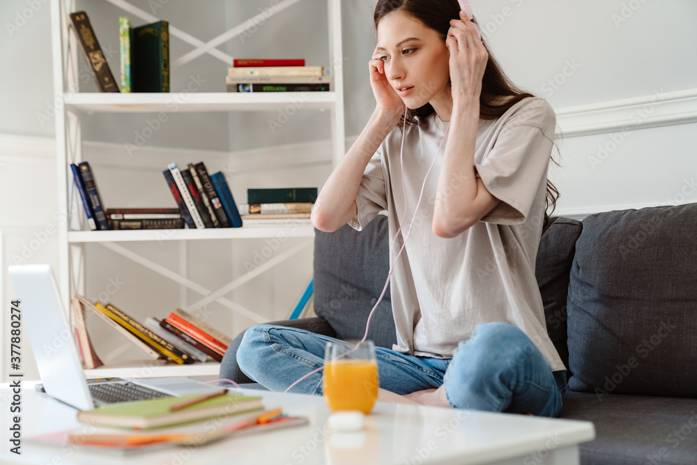 Image of serious girl using laptop and headphones while sitting on sofa Image of serious girl using laptop and headphones while sitting on sofa