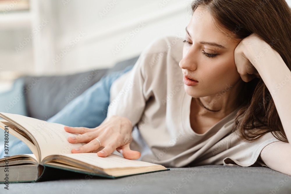 Image of concentrated beautiful girl reading book while lying on sofa Image of concentrated beautiful girl reading book while lying on sofa