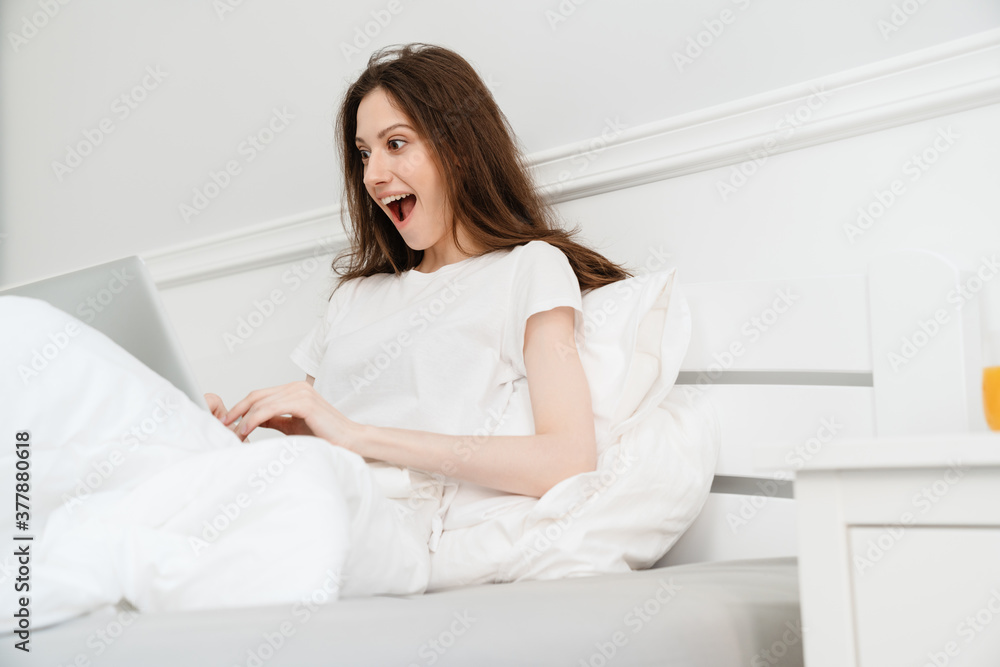 Image of surprised beautiful girl using laptop while sitting in bed Image of surprised beautiful girl using laptop while sitting in bed