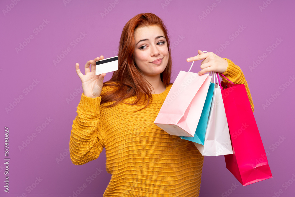 Redhead teenager girl over isolated purple background holding shopping bags and a credit card Redhead teenager girl over isolated purple background holding shopping bags and a credit card