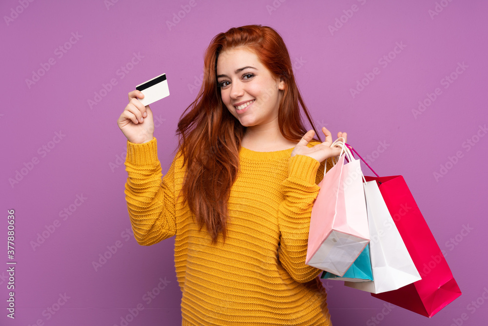Redhead teenager girl over isolated purple background holding shopping bags and a credit card Redhead teenager girl over isolated purple background holding shopping bags and a credit card