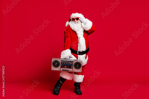 Full length photo of pensioner old man grey beard funny posing hold glasses boom box wear santa costume gloves coat belt sunglass headwear black boots isolated red color background