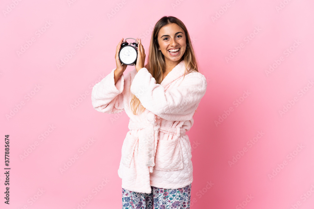 Young hispanic woman over isolated pink background in pajamas and holding clock with happy expression Young hispanic woman over isolated pink background in pajamas and holding clock with happy expression
