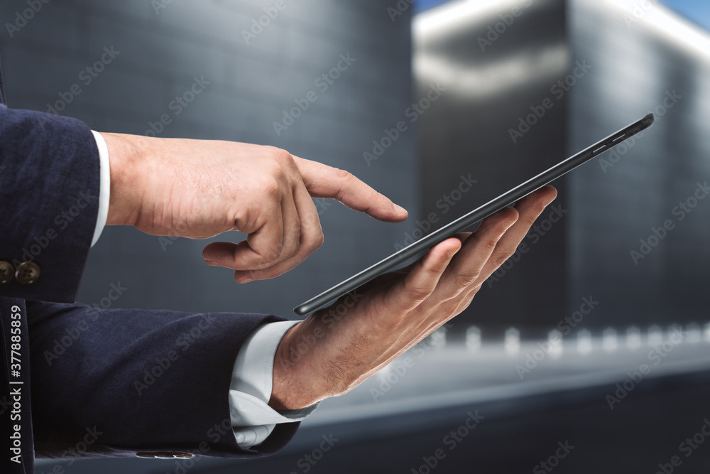 Businessman hand clicks on the screen of a digital tablet near the exterior of the business center at night, close up. Internet Work Concept