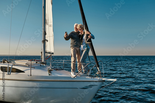 Happy moments. Full length of beautiful senior couple making selfie on smartphone and smiling while standing together on the side of sailboat or yacht deck floating in sea © Kostiantyn