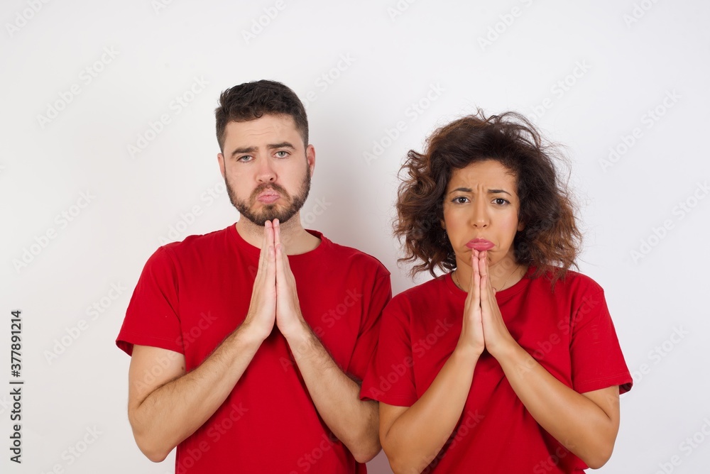 Positive Young beautiful couple wearing red t-shirt on white background smiles happily, glad to receive pleasant news from interlocutor, keeps palms together, People emotions concept. Positive Young beautiful couple wearing red t-shirt on white background smiles happily, glad to receive pleasant news from interlocutor, keeps palms together, People emotions concept.