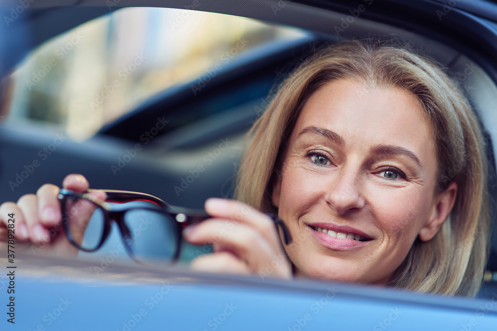 Portrait of a happy business woman holding eyeglasses and smiling at camera while sitting behind steering wheel of her modern car, driving through the city