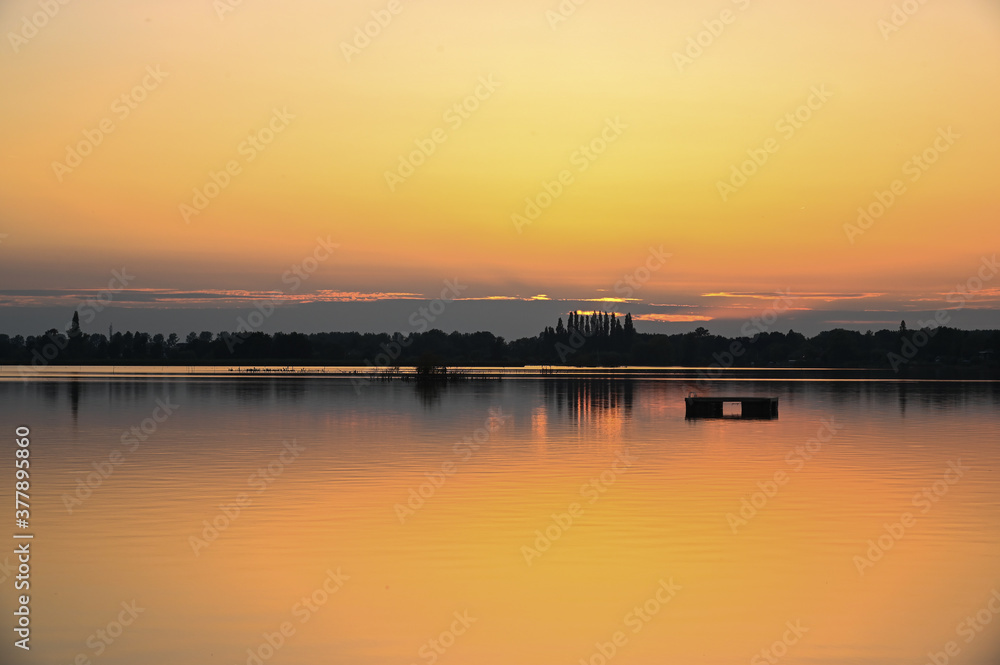 Horizon with the silhouettes of trees and shrubs surmounted by an orange sunset that is reflected in the water of a lake. Reeuwijk, The Netherlands, Europe.