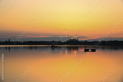 Horizon with the silhouettes of trees and shrubs surmounted by an orange sunset that is reflected in the water of a lake. Reeuwijk, The Netherlands, Europe. © Karin Reine