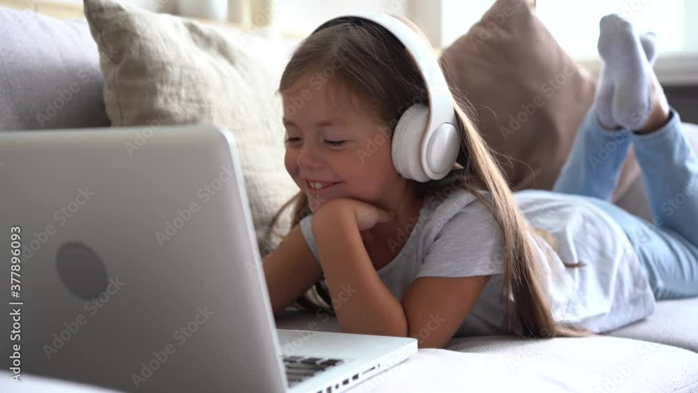 Smiling school girl wearing headphones using laptop lying on sofa, communicating with friend by webcam at home Smiling school girl wearing headphones using laptop lying on sofa, communicating with friend by webcam at home