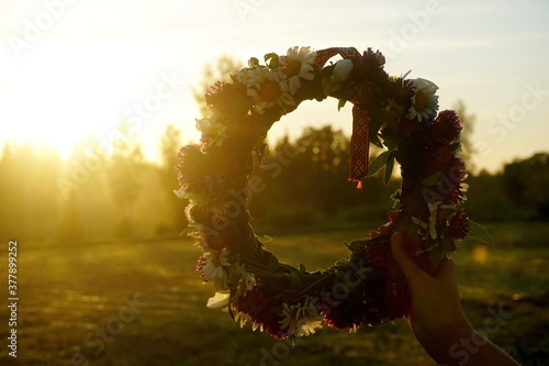 Female hands holding a wreath of wild flowers and a red and white ribbon with signs on a background of evening sunset