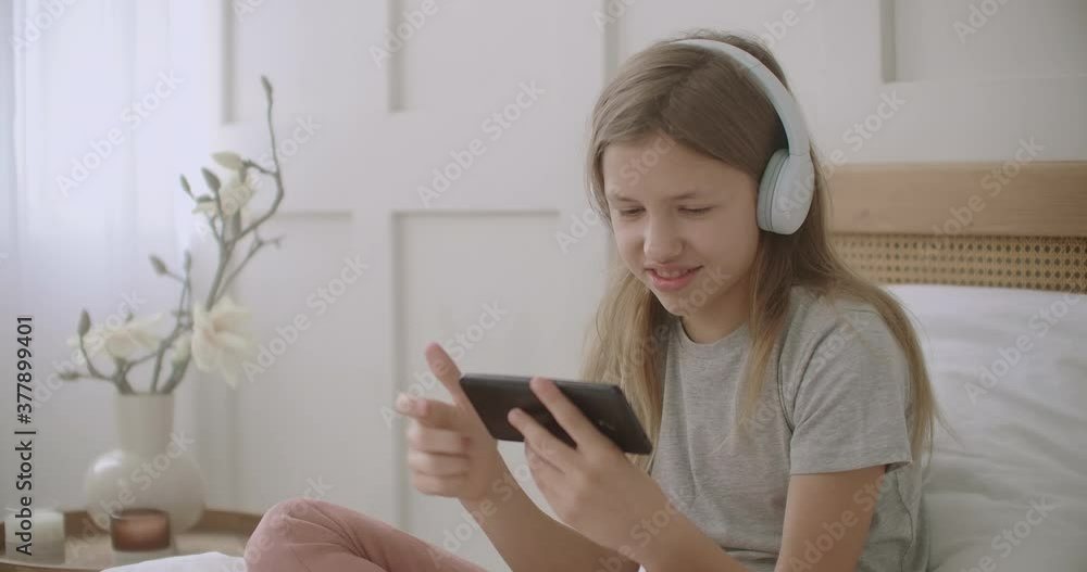 little girl is using smartphone and wireless headphones sitting on bed in room, playing game and smiling little girl is using smartphone and wireless headphones sitting on bed in room, playing game and smiling