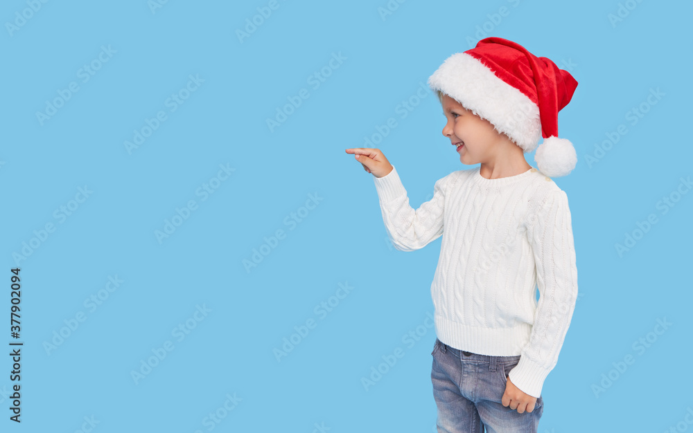 Portrait of a satisfied little child boy dressed in christmas Santa hat. laughing isolated over blue background. Pointing hand to the left Portrait of a satisfied little child boy dressed in christmas Santa hat. laughing isolated over blue background. Pointing hand to the left