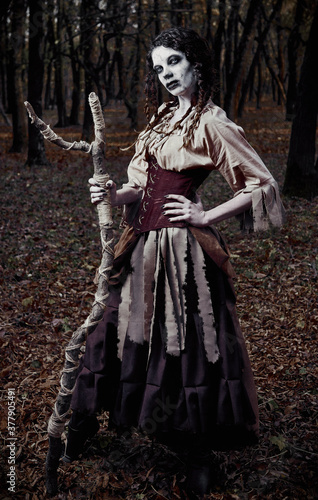 Halloween theme: ugly scary voodoo witch with stick. Portrait of evil sorceress in dark forest. Zombie woman (undead)