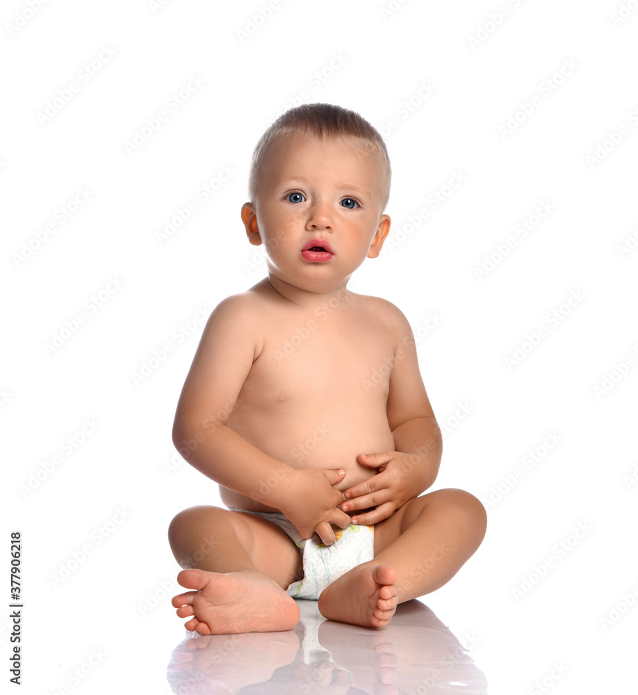 Calm and curious baby boy toddler naked in diaper sits on the floor, touches his stomach and looks at camera Calm and curious baby boy toddler naked in diaper sits on the floor, touches his stomach and looks at camera