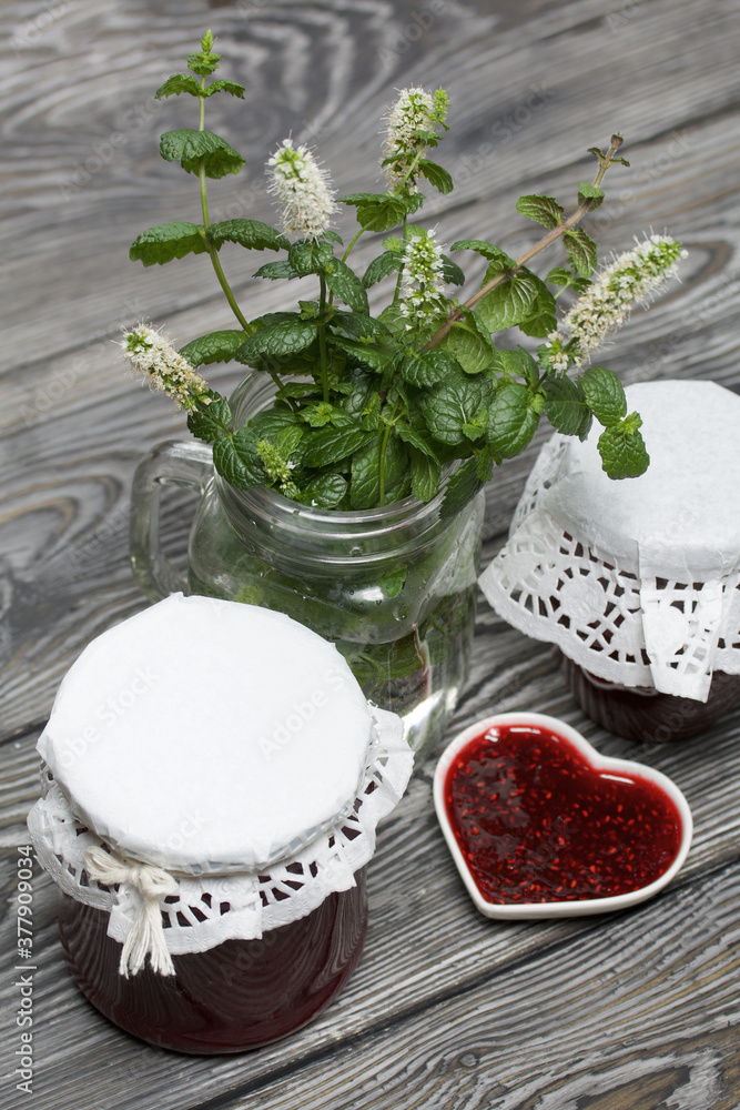 Raspberry jam in jars and a saucer. On pine planks painted black and white. Nearby flowering branches of mint in a jar.