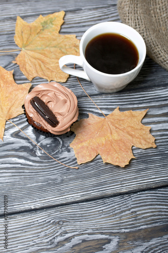 Chocolate cupcakes with cream cheese cream. Nearby is a cup of coffee and dried maple leaves. On pine planks painted black and white.