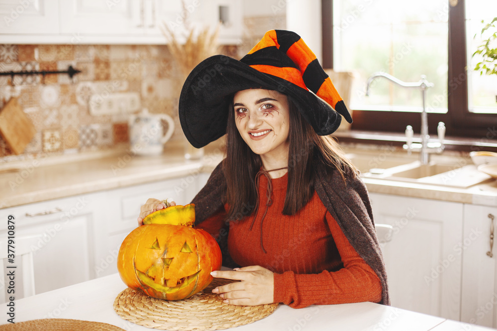 Happy and beautiful girl in the kitchen at home in a suit and make-up with a pumpkin lantern smiling Happy and beautiful girl in the kitchen at home in a suit and make-up with a pumpkin lantern smiling