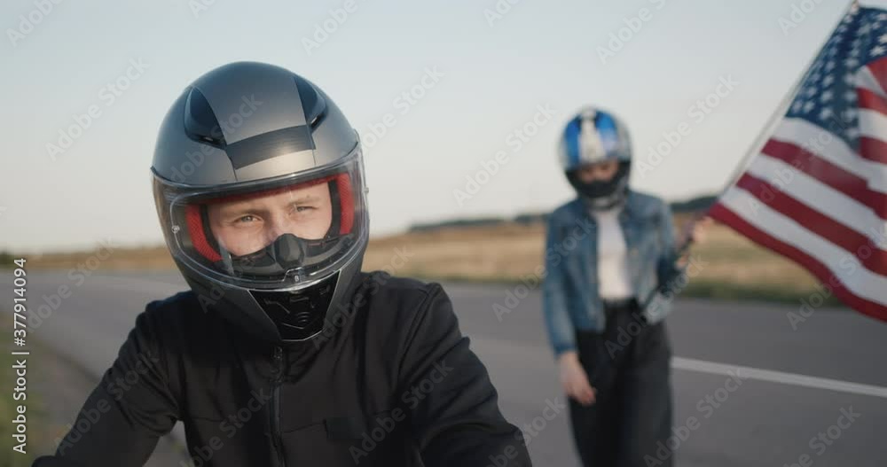 A young female biker in a helmet with a U.S. flag sits down to his boyfriend on a motorcycle A young female biker in a helmet with a U.S. flag sits down to his boyfriend on a motorcycle