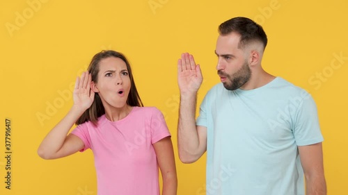 Young fun couple friends woman man 20s in basic t-shirts isolated on yellow background studio. Look aside hold hand near ear try to hear you overhear listening intently sharing news whispering secret photo