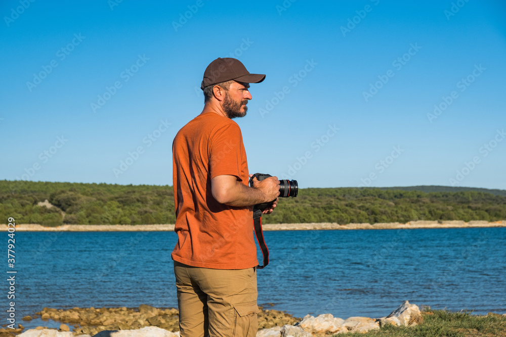 Obraz premium Photographer man in t-shirt and cargo pants walking around sea shore with DSLR camera and shooting nature, half body portrait, landscape photography concept