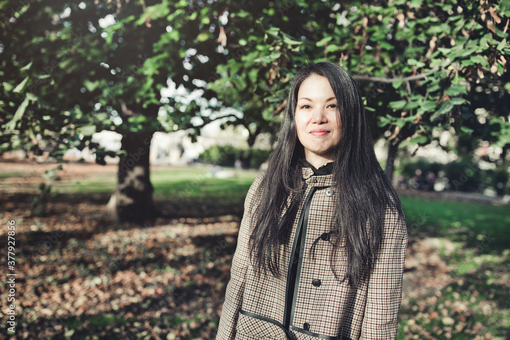 Beautiful middle-aged Japanese woman in stylish coat in autumn park, smiling. Sunlight, copy space Beautiful middle-aged Japanese woman in stylish coat in autumn park, smiling. Sunlight, copy space
