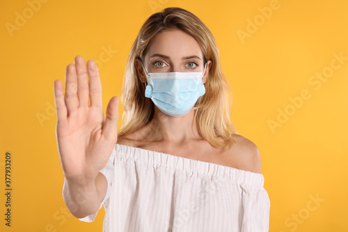 Woman in protective face mask showing stop gesture on yellow background. Prevent spreading of coronavirus © New Africa