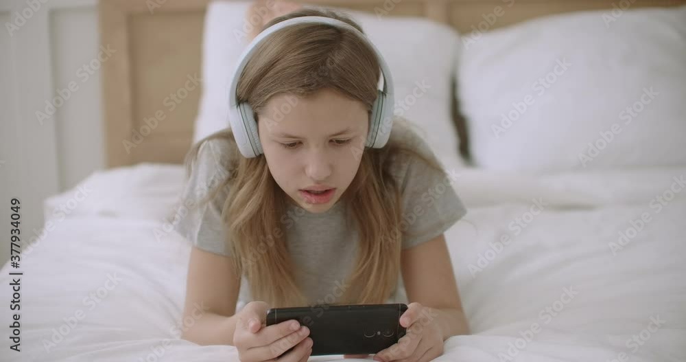 joyful teen girl is reading text on screen of mobile phone, lying on bed, using wireless headphones and wifi joyful teen girl is reading text on screen of mobile phone, lying on bed, using wireless headphones and wifi