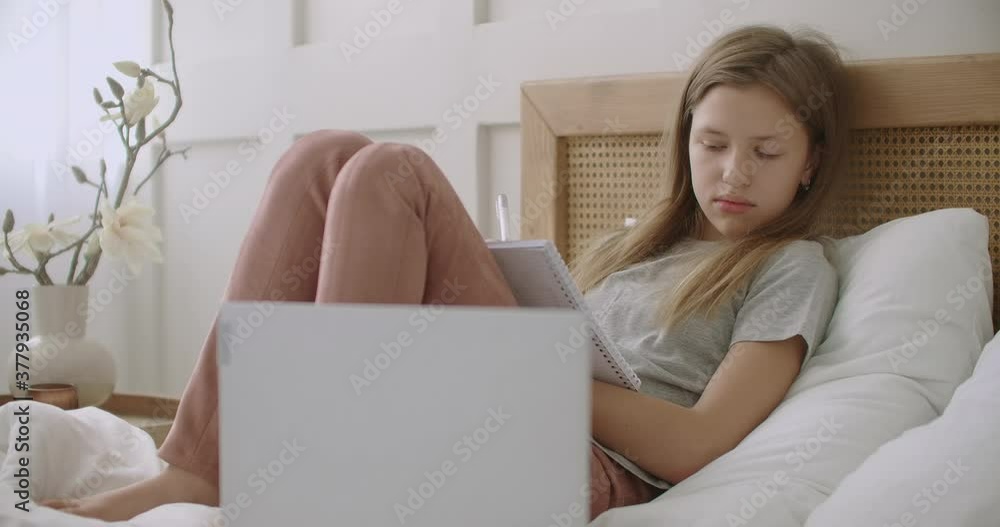 tired and bored pupil girl is lying on bed with exercise book and listening lesson on laptop, e-learning for school children tired and bored pupil girl is lying on bed with exercise book and listening lesson on laptop, e-learning for school children