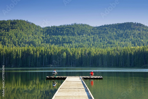 Original outdoor photograph of the reflection of two fisherman fishing on opposites ends of a long dock on a lake 