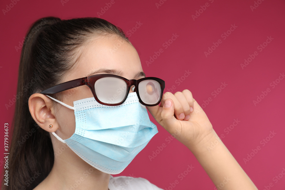 Little girl wiping foggy glasses caused by wearing medical face mask on pink background. Protective measure during coronavirus pandemic Little girl wiping foggy glasses caused by wearing medical face mask on pink background. Protective measure during coronavirus pandemic