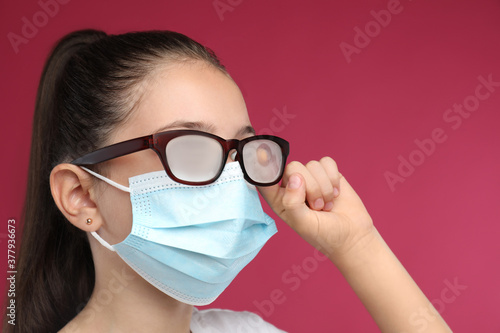 Little girl wiping foggy glasses caused by wearing medical face mask on pink background. Protective measure during coronavirus pandemic