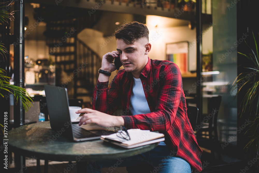Focused male freelancer using laptop and talking on smartphone in cafe Focused male freelancer using laptop and talking on smartphone in cafe