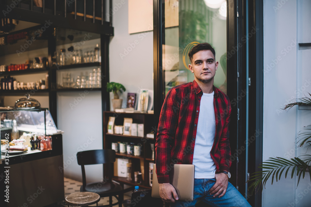 Confident man standing near entrance of cafe with laptop Confident man standing near entrance of cafe with laptop