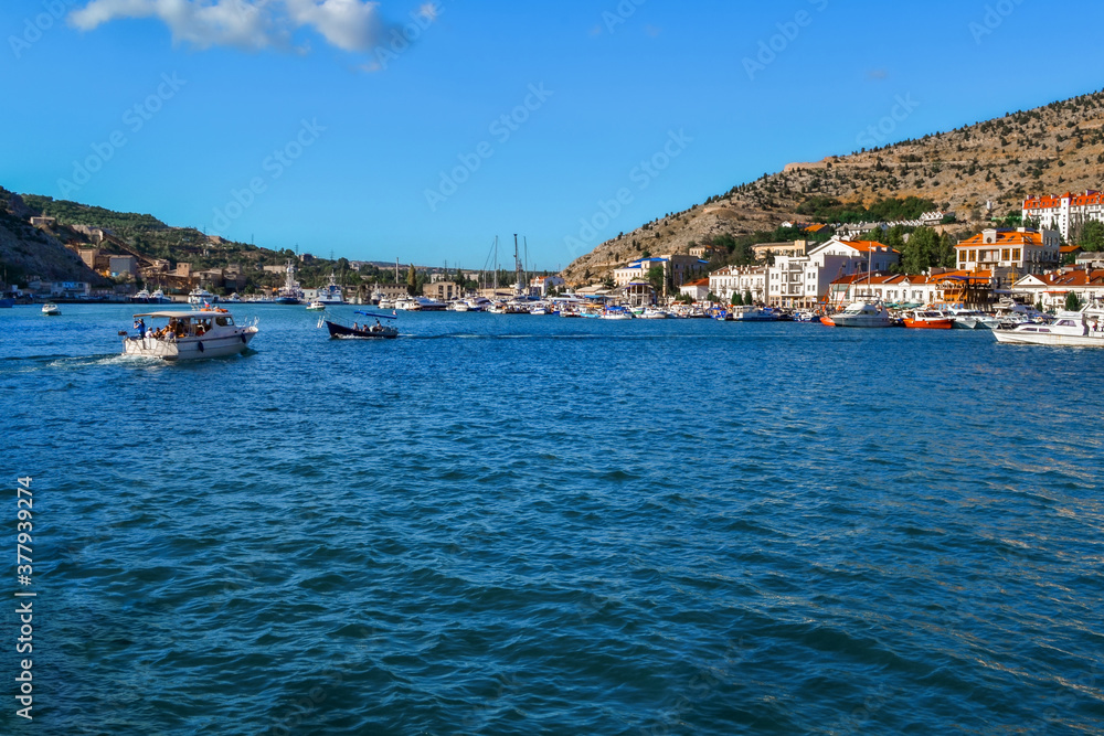 blue water with waves in bay of mountain town of Balaklava with marinas, white ships, boats and bright buildings, ripples, summer