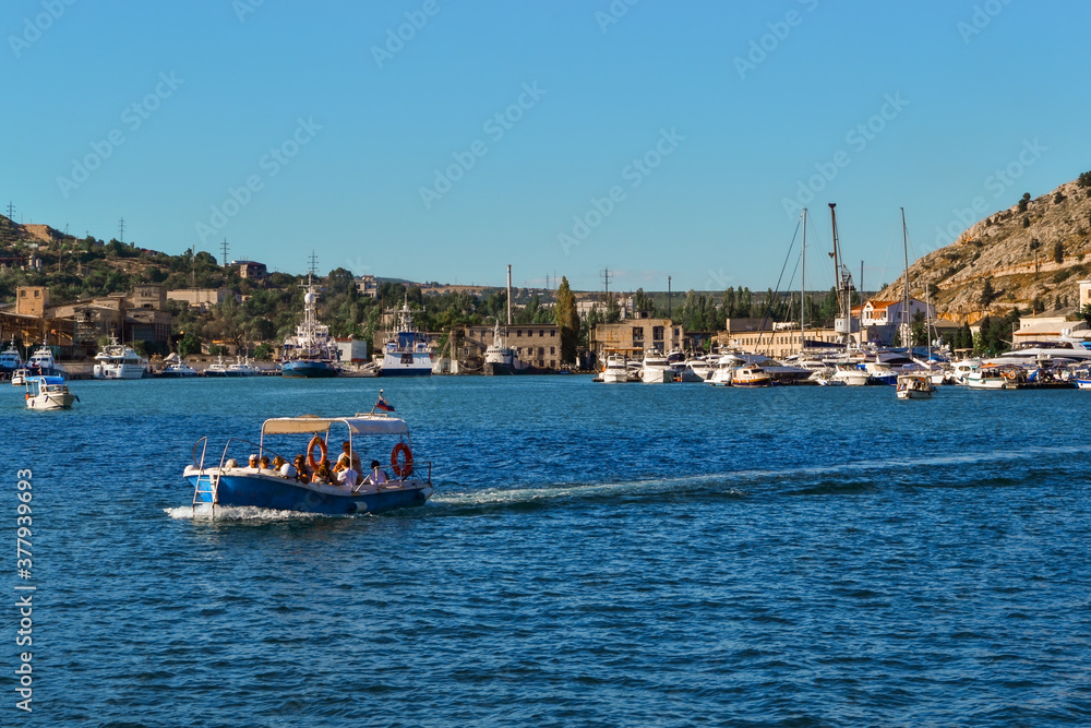 bright blue boat with people sails along the bay of resort town of Balaklava among white ships on docks near houses and buildings, sunny