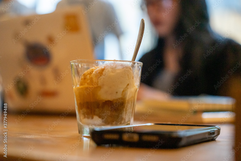 Cold Brew Iced Coffee Affogato, Vanilla Ice-Cream in a glass next to smart phone on wooden table and customers using laptop for work in blurred background. Cold Brew Iced Coffee Affogato, Vanilla Ice-Cream in a glass next to smart phone on wooden table and customers using laptop for work in blurred background.
