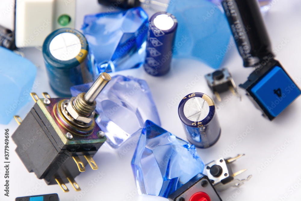 Closeups of capacitors and switches set against a white background with blue marbles and crystals in the foreground. Closeups of capacitors and switches set against a white background with blue marbles and crystals in the foreground.