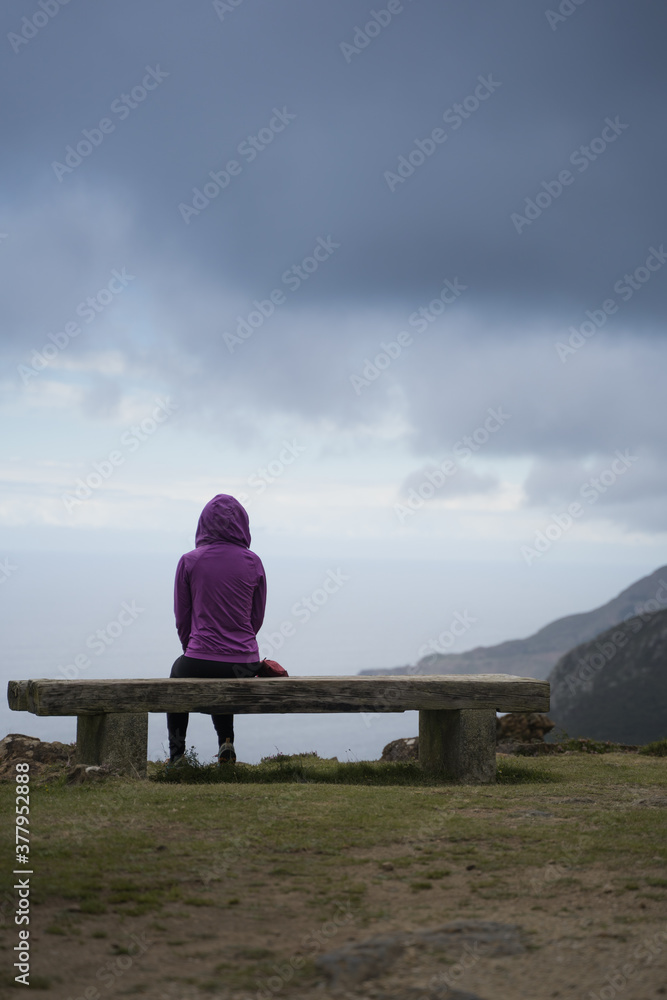 Thoughtful Hooded person is Looking at Ocean siting on a stone bench on top of a high cliff. Back view of woman contemplating the Sea and the Stormy Clouds