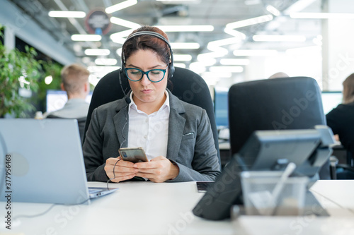 Business woman dressed in a headset is bored and uses a smartphone while sitting at a desk. Female manager is distracted from work by phone