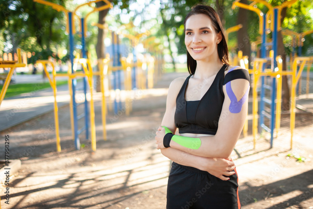 Portrait of attractive muscular brunette woman wearing black sports outfit, looking at camera. Young smiling female athlete posing with arms crossed, colorful kinesiotaping on body.