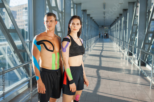 Portrait of muscular brunette woman with arms crossed and man looking at camera with strong face. Young couple athletes posing indoors, colorful kinesiotaping on body, futuristic interior.