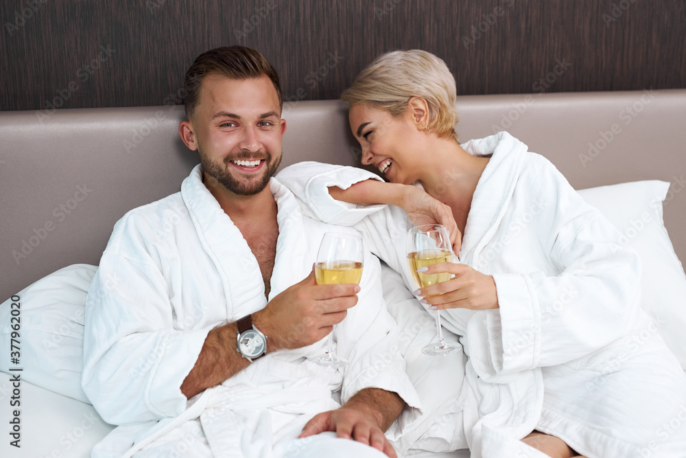 happy caucasian couple have romantic time in hotel, young man and woman lie on bed in white bathrobes and drink champagne, relax