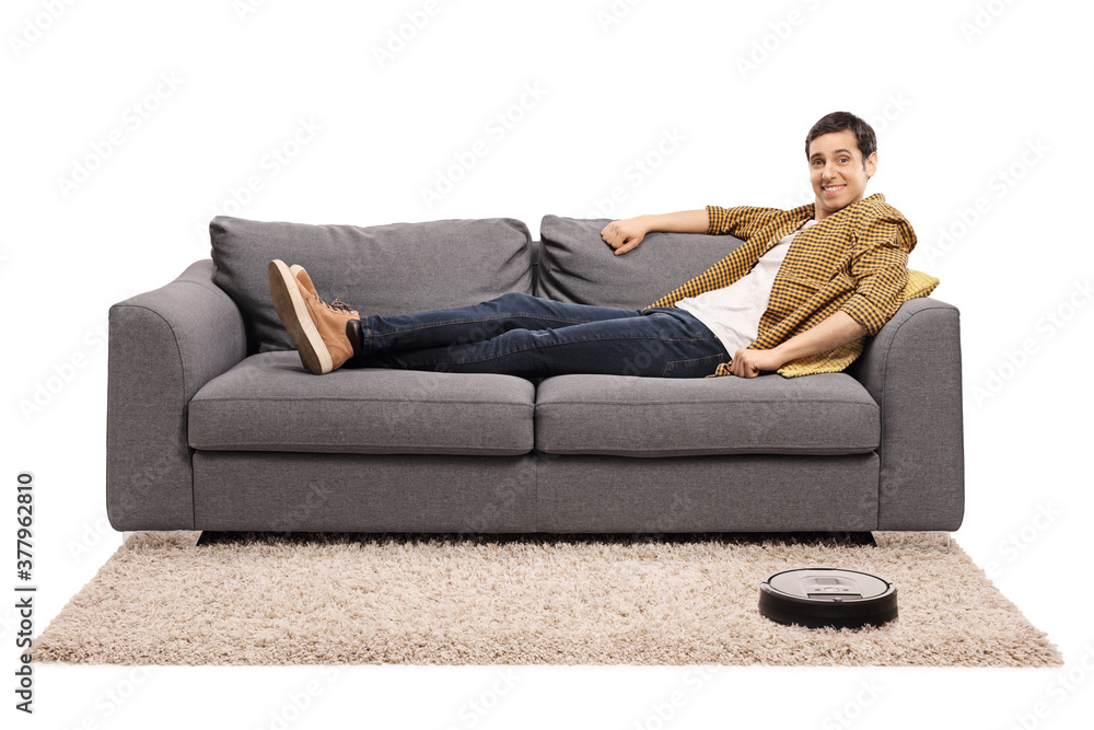 Young man sitting on a sofa and a robotic vacuum cleaner vacuuming the carpet Young man sitting on a sofa and a robotic vacuum cleaner vacuuming the carpet