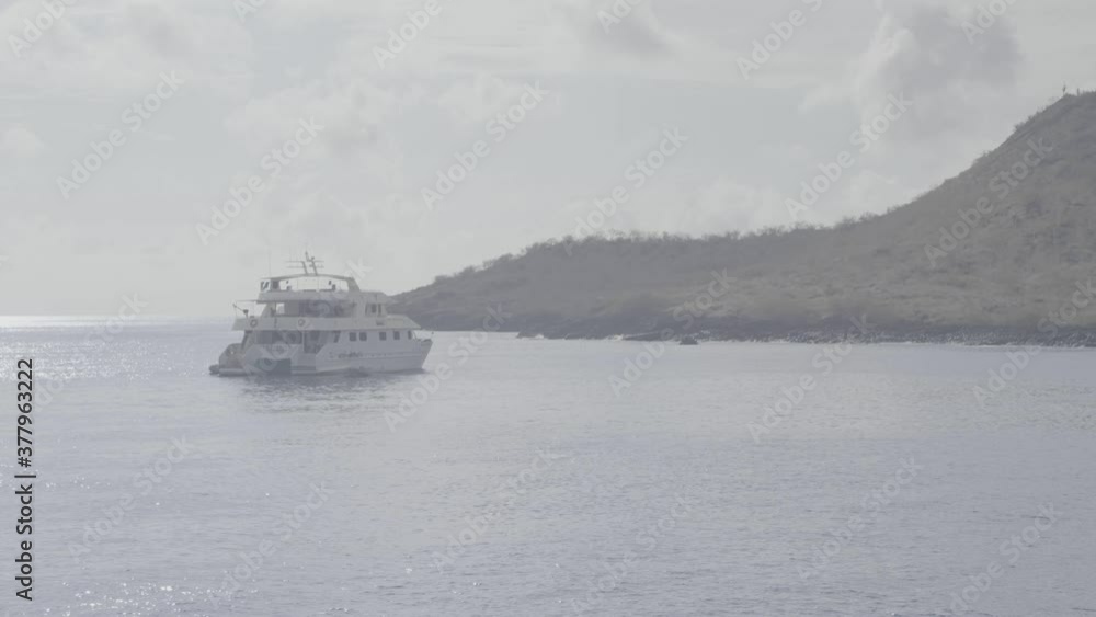 Near an island of the Galapagos islands, a small white cruise and yacht is anchored on a calm ocean. Near an island of the Galapagos islands, a small white cruise and yacht is anchored on a calm ocean.