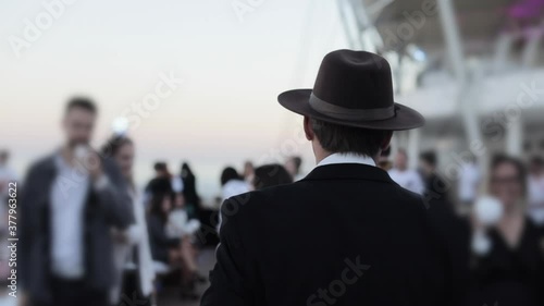 A man with a black hat on his head walks among other people at the beach club. The camera captures the man from the back.